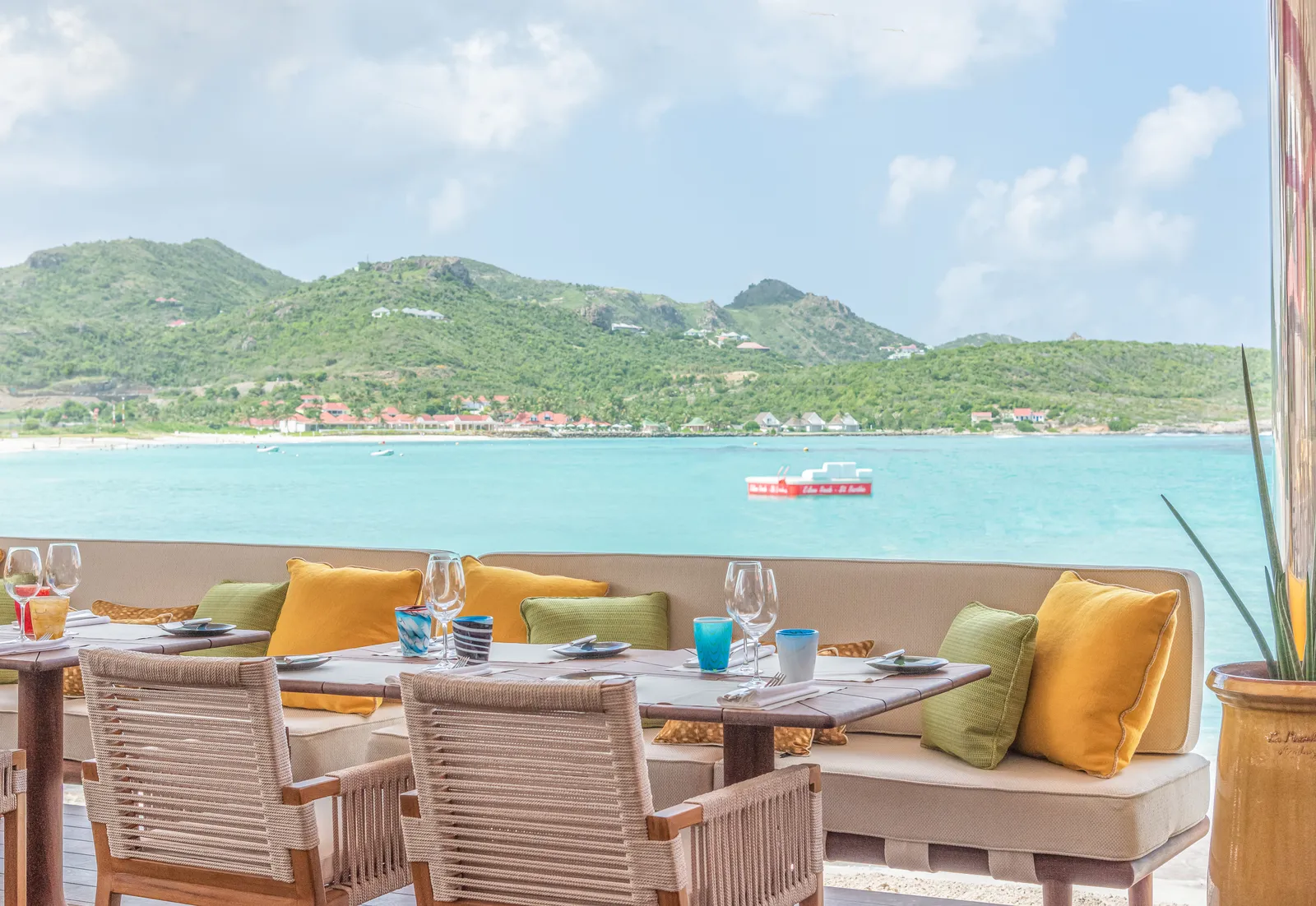 A laid table at the Sand Bar with a bench of yellow cushions, facing the bay and the green hills.