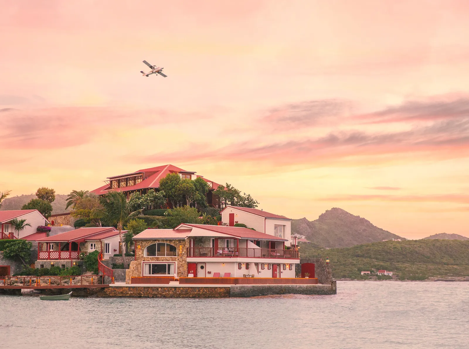 The rock and its red roofs at dawn under an orange and pink sky, with a small aeroplane crossing above the bay.