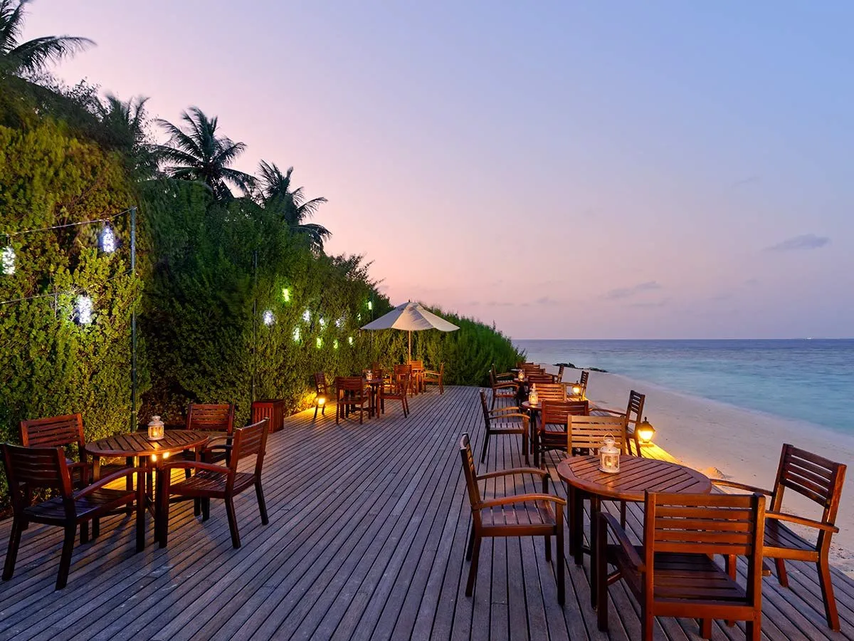 The bar’s timber deck over the sea at dusk, with its tables, lit lanterns and palms.