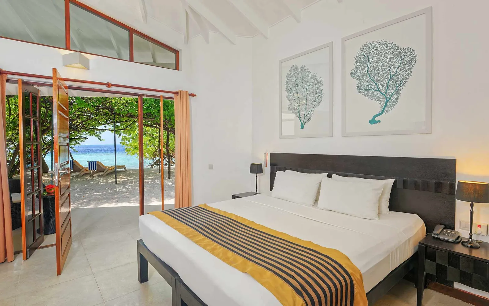A bungalow bedroom: the double bed, coral prints on the walls and the doors open onto the beach.
