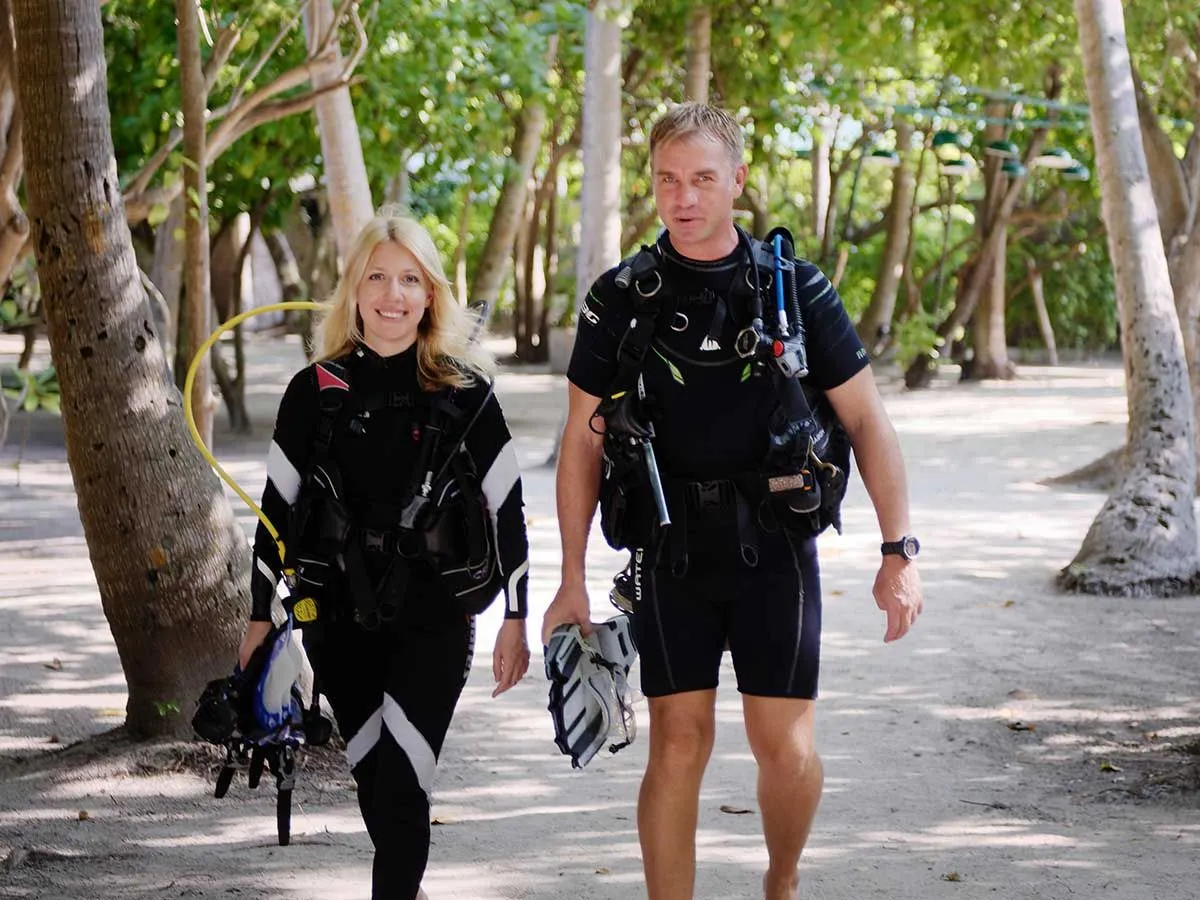 2 divers in wetsuits and jackets walk across the sand between the palms, fins in hand.