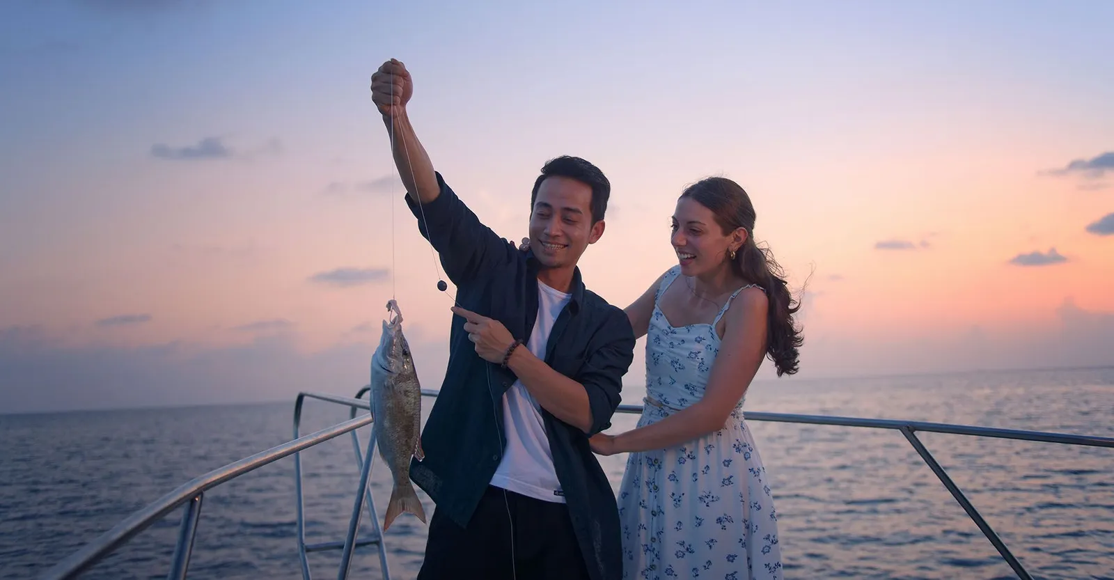 A couple at the bow of a boat at sunset: he holds up a fish just caught on a hand line, she laughs.
