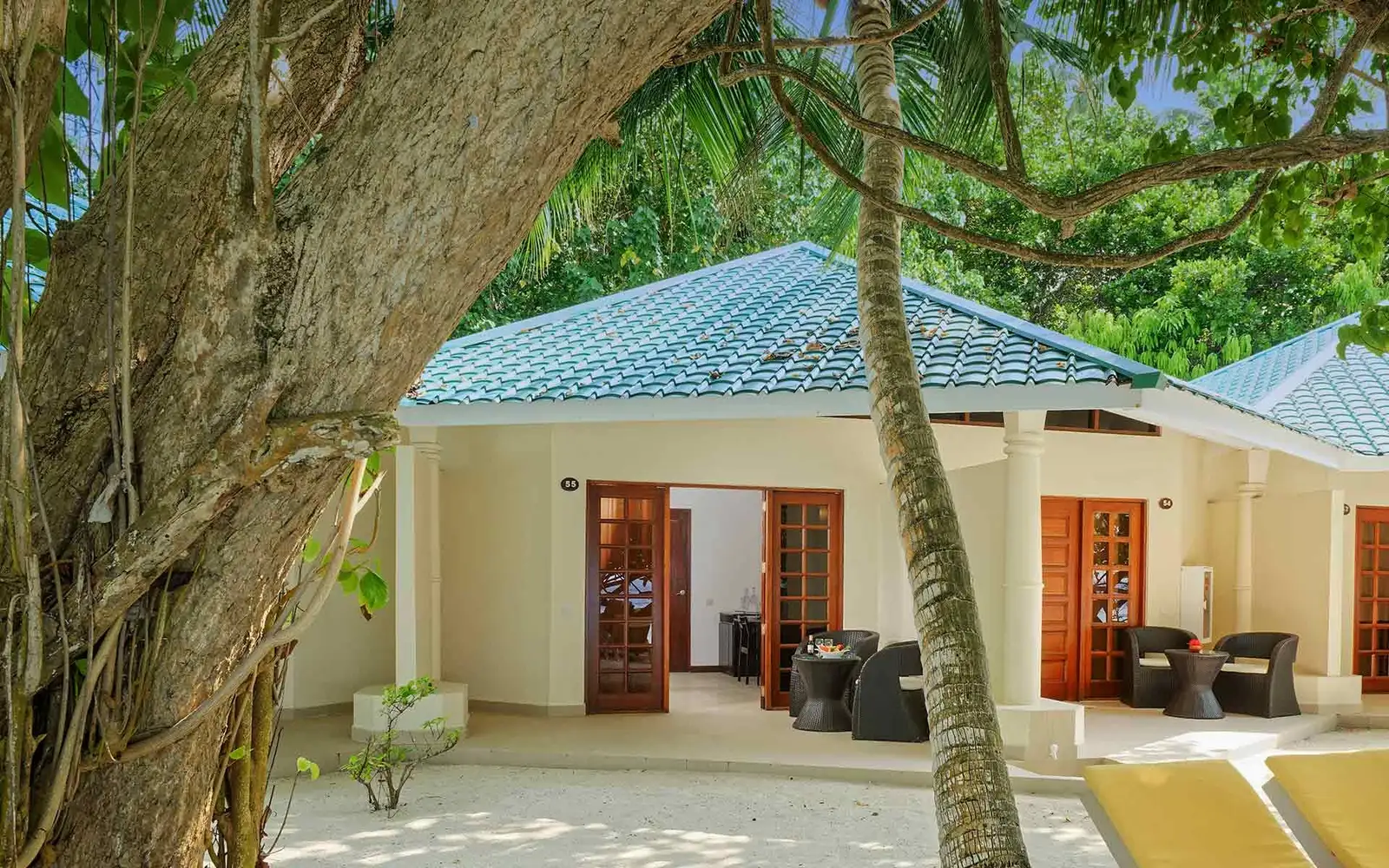 A beach bungalow with a blue roof, its timber doors open and armchairs under the porch, in the shade of the trees.