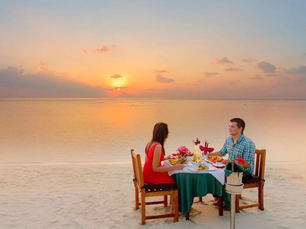 A couple dine at a table laid on the sand, facing the sun setting over the sea.