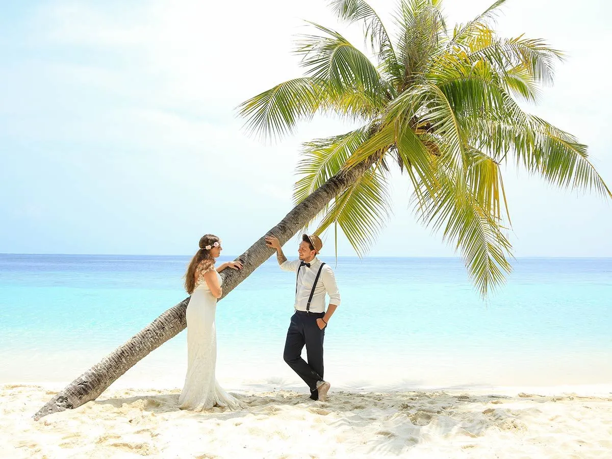 A couple dressed for a wedding beneath a leaning palm on the beach, facing the sea.
