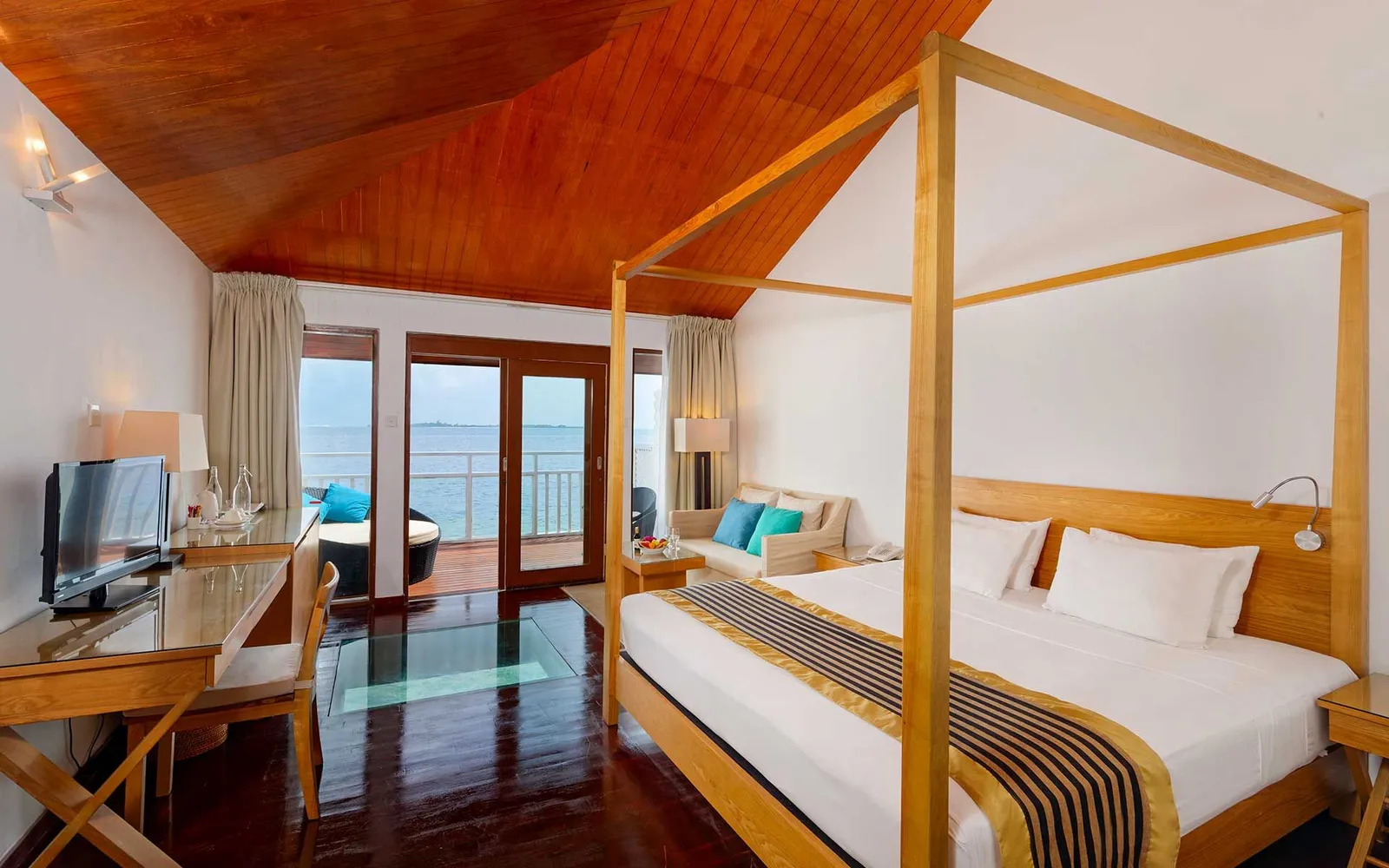 A water villa bedroom: the pale four-poster bed, the red ceiling and the glass doors onto the balcony.