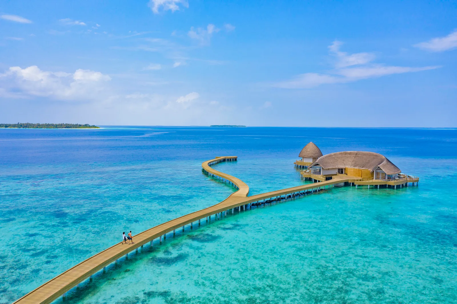 The timber jetty crossing the turquoise lagoon to the thatched roofs of Mediterraneo and Eclipse, with 2 people walking along it.