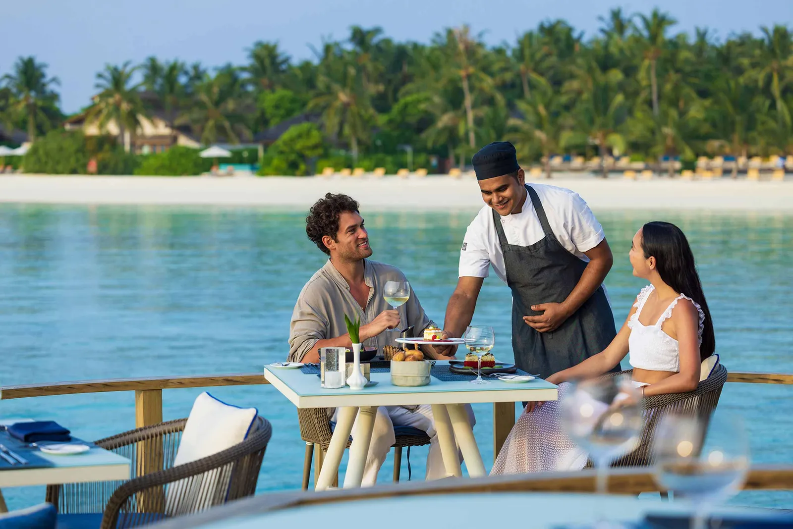 A couple at a table on Mediterraneo’s deck above the clear water, as a cook in an apron brings the dishes.