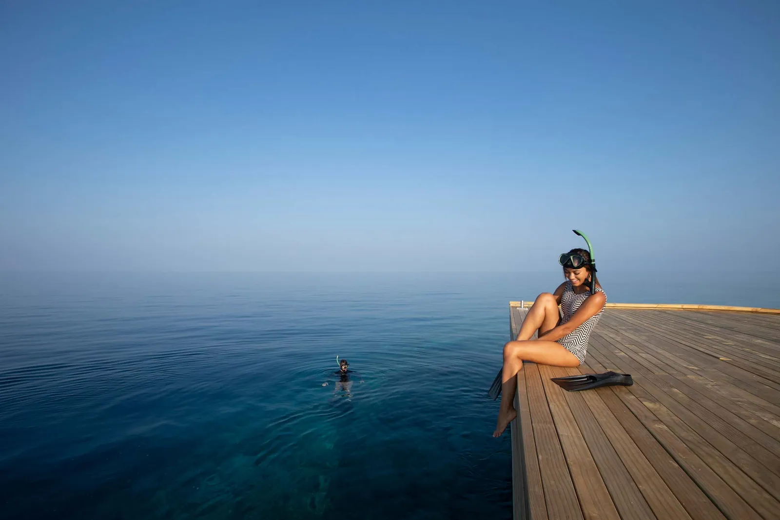 A young woman in a mask sitting on the edge of a timber deck over the open sea, a swimmer in the water below.