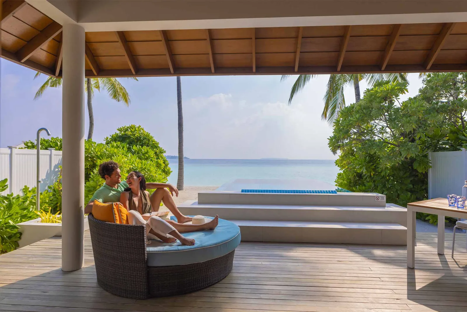 A couple on the round daybed of a Beach Villa with Pool’s covered deck, the pool and the sea in front.