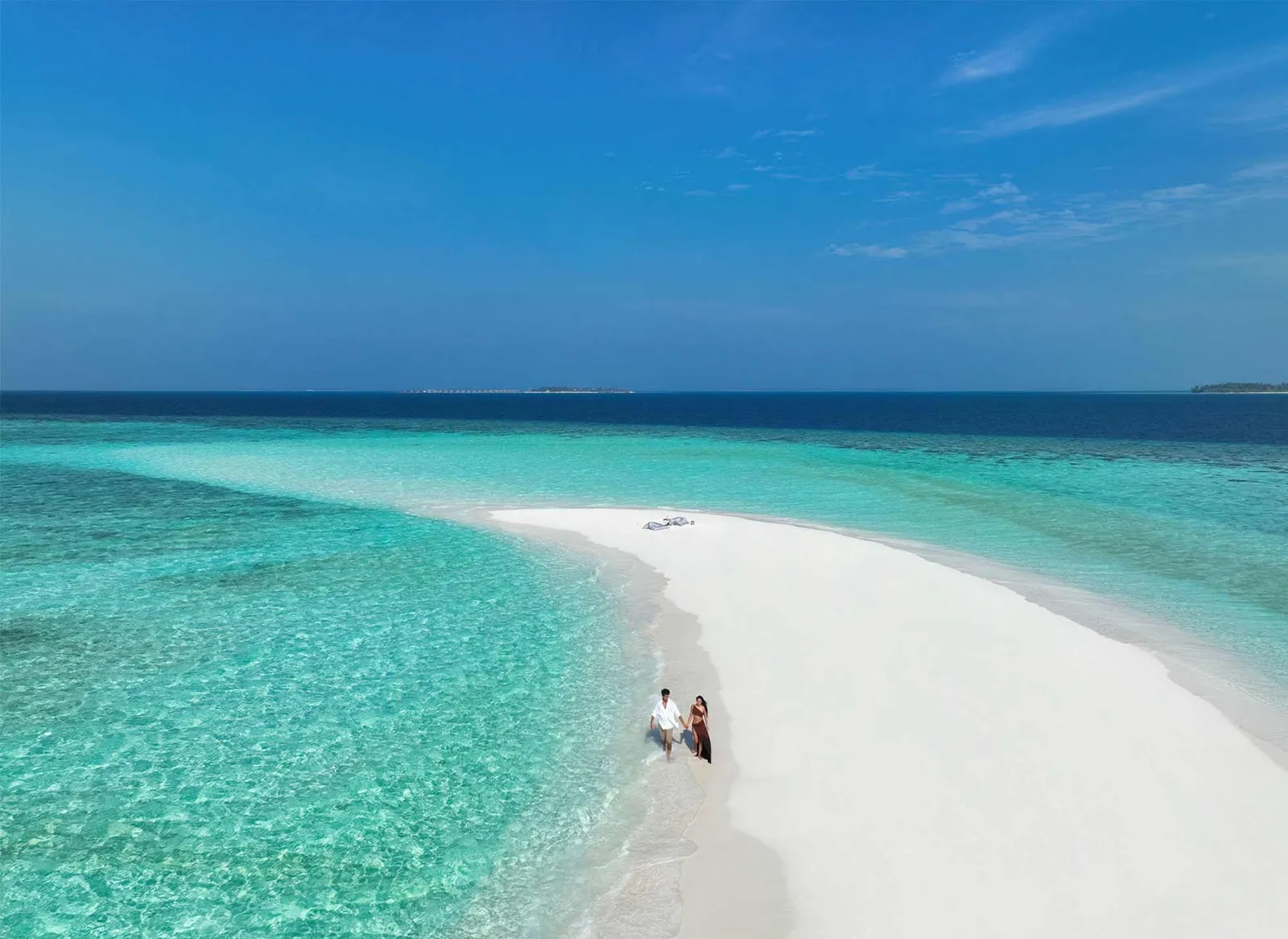 A couple at the end of a long spit of white sand, turquoise water all around.