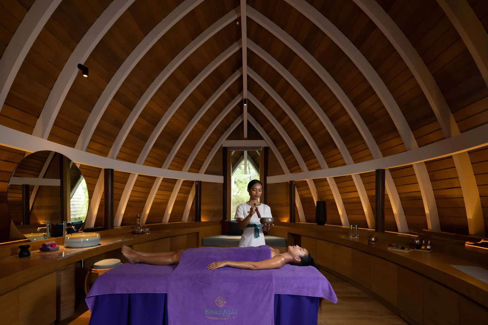 An Emerald Spa room under its timber ribs, a guest lying on the purple couch and the therapist standing behind.