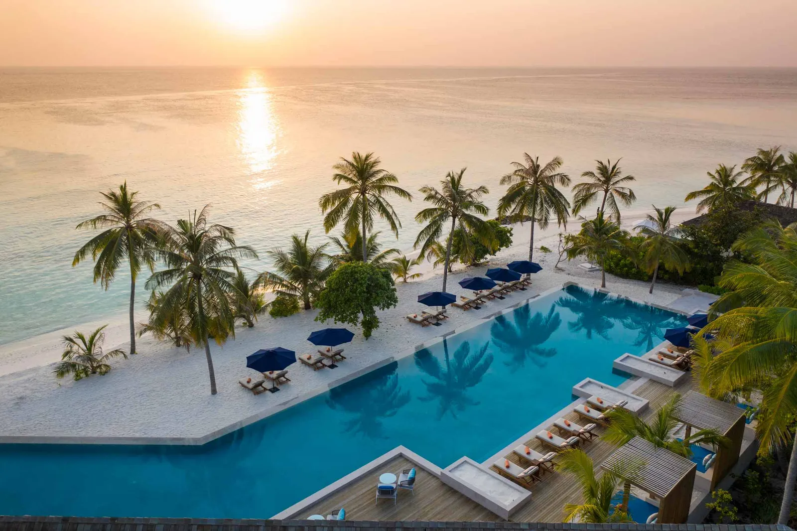The Sunset Pool & Bar at sunset from above, with loungers, blue parasols and palms along the beach.
