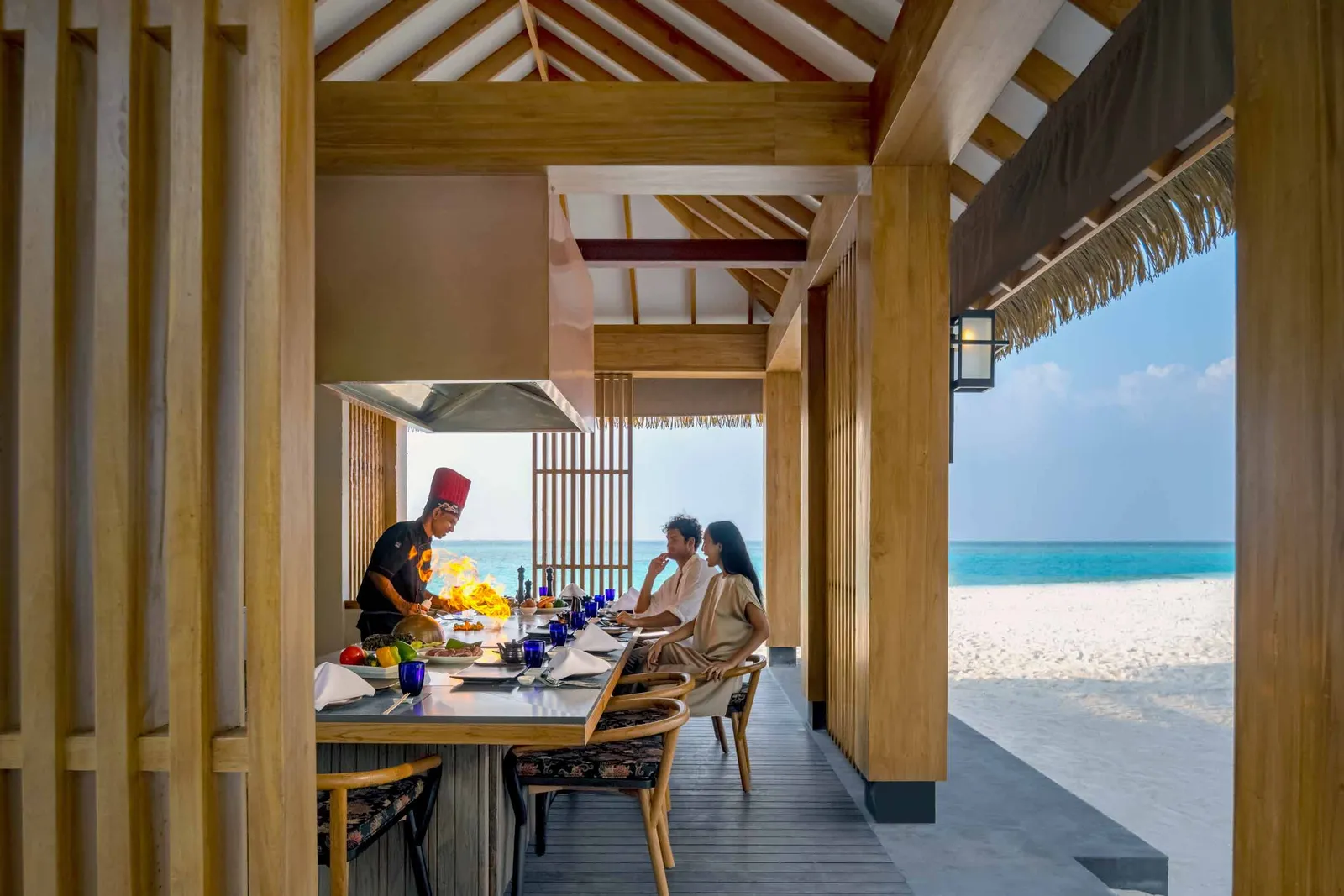 The teppanyaki counter: the chef in a red hat at the hotplate in front of 2 guests, the beach beyond the pillars.