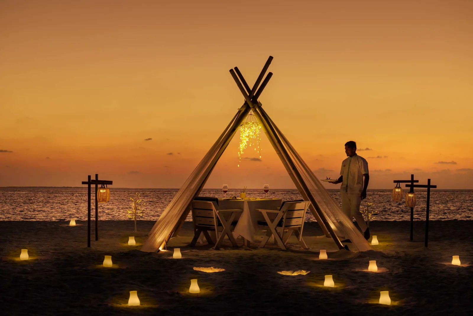 A dinner for 2 on the beach at sunset, under a pointed canopy with lanterns lit on the sand.