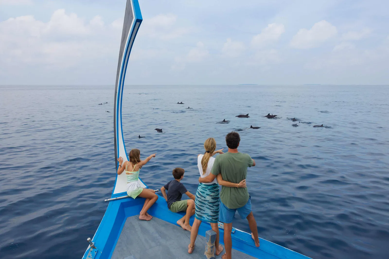 A family at the bow of a dhoni watching a pod of dolphins swim across the calm sea.