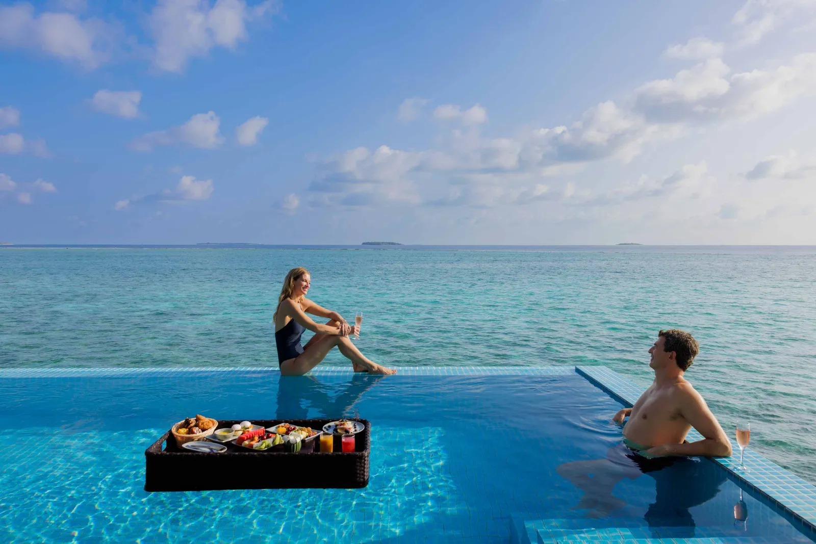 A couple in a water villa’s infinity pool, a breakfast tray floating in front of them and the sea beyond.