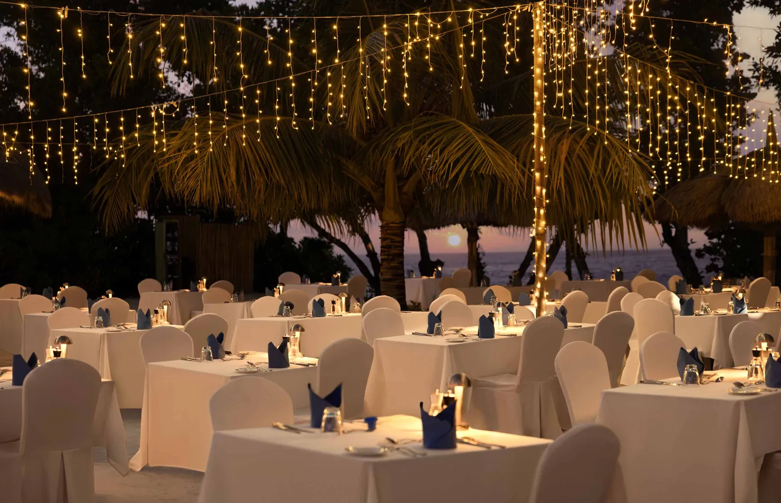 A gala dinner on the beach at night, tables laid in white under lights strung from the trees.
