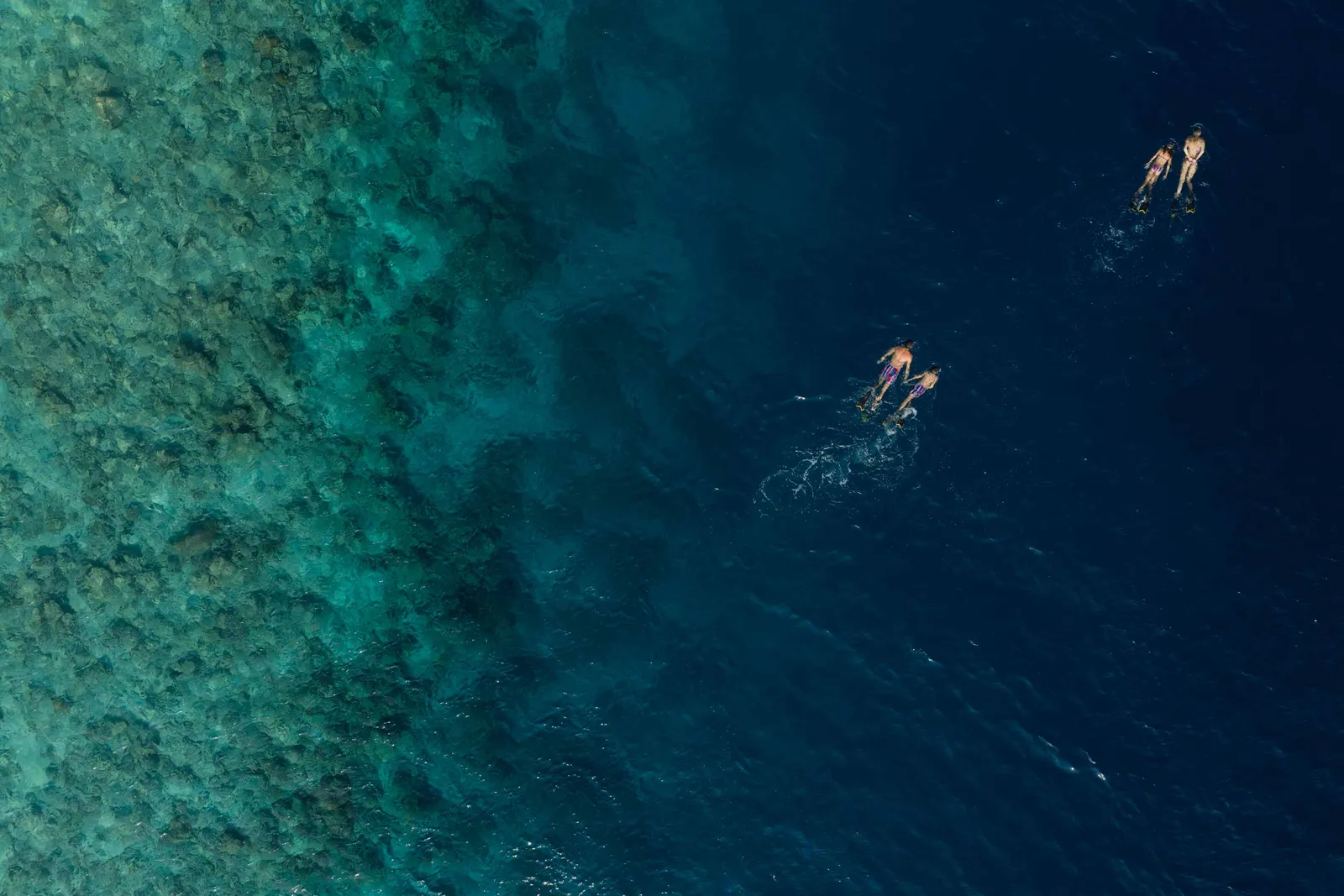 2 people snorkelling at the edge of the lagoon, seen from above, where the pale water turns blue.
