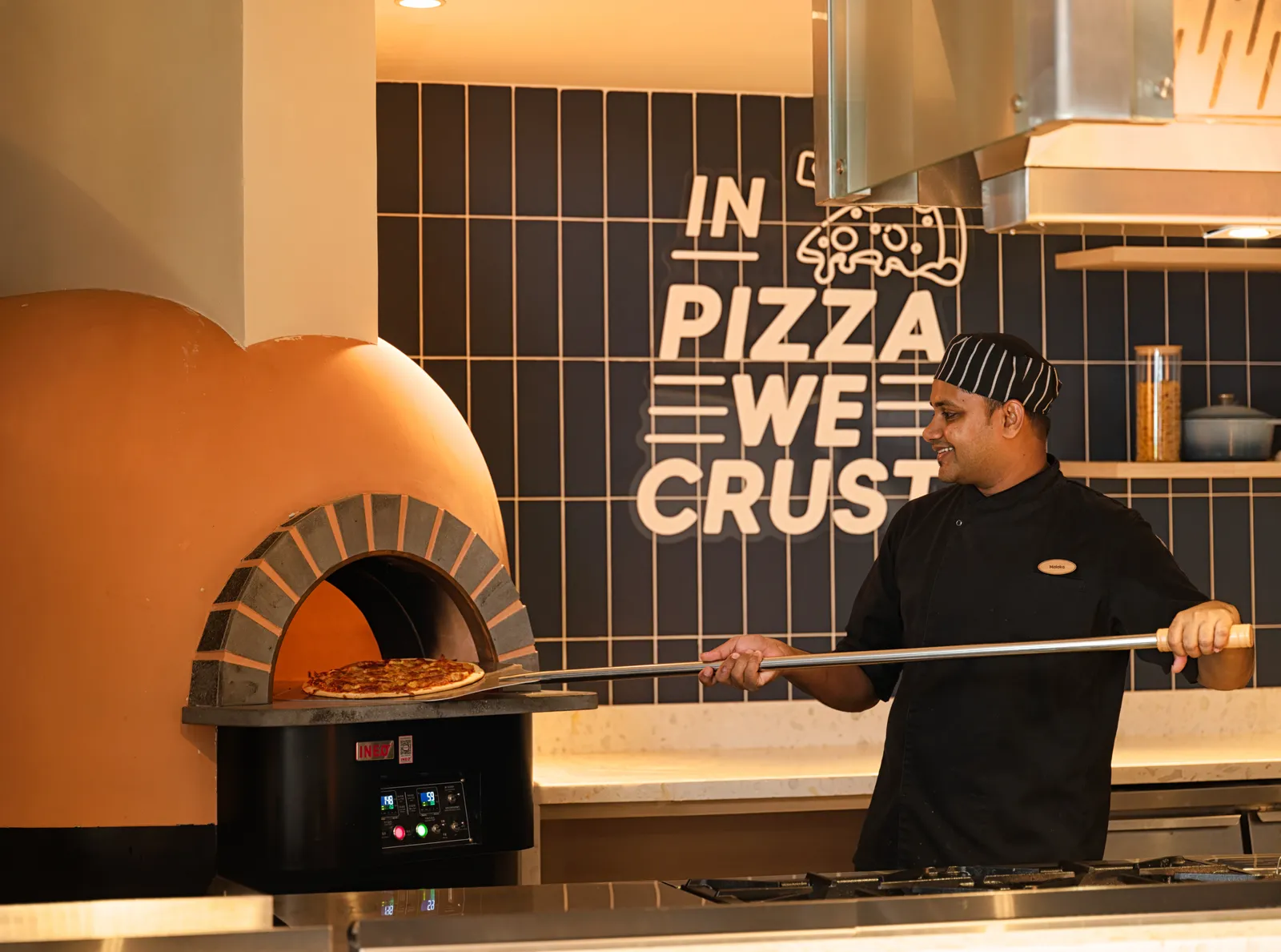 A chef slides a pizza into Soul Kitchen’s wood-fired oven, under the words «In pizza we crust».