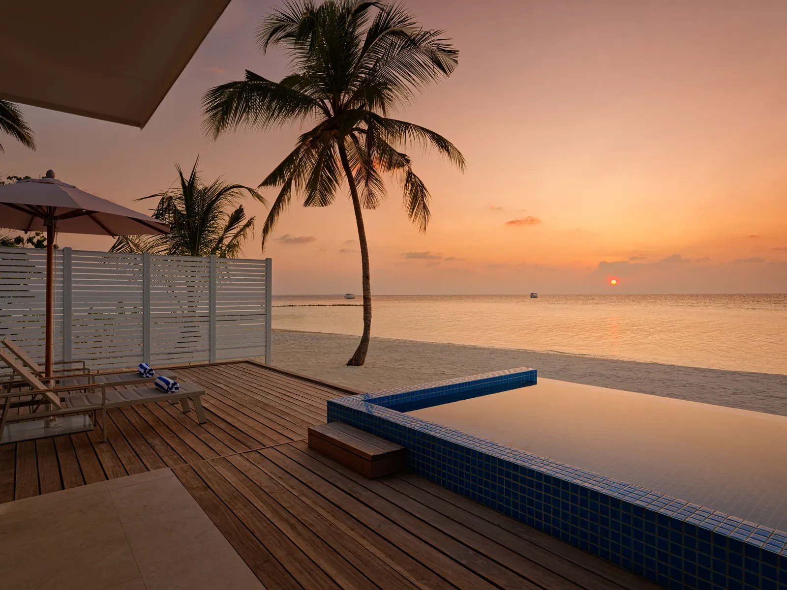 The timber deck of a Beach Pool Villa at sunset, with its private pool, a palm and a pink sea.
