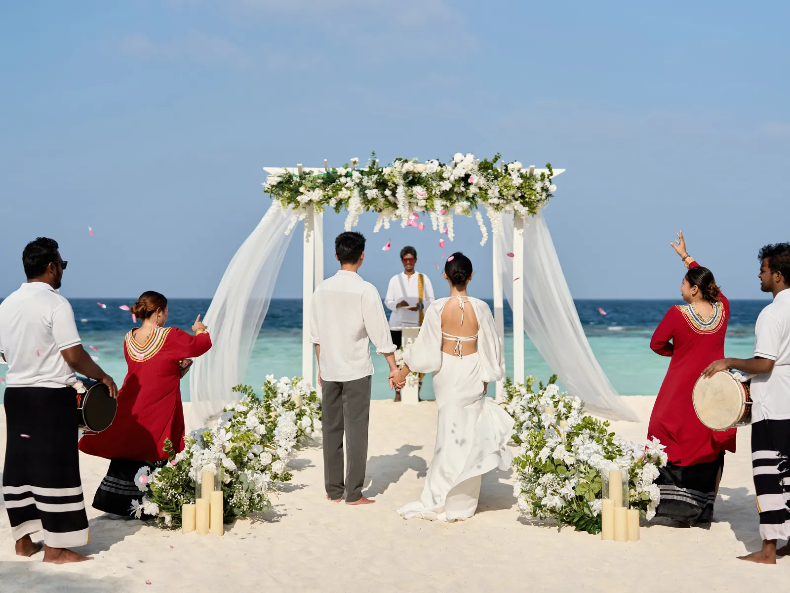 A couple under a floral arch on the beach, among bodu beru drummers and 2 women in red dresses.