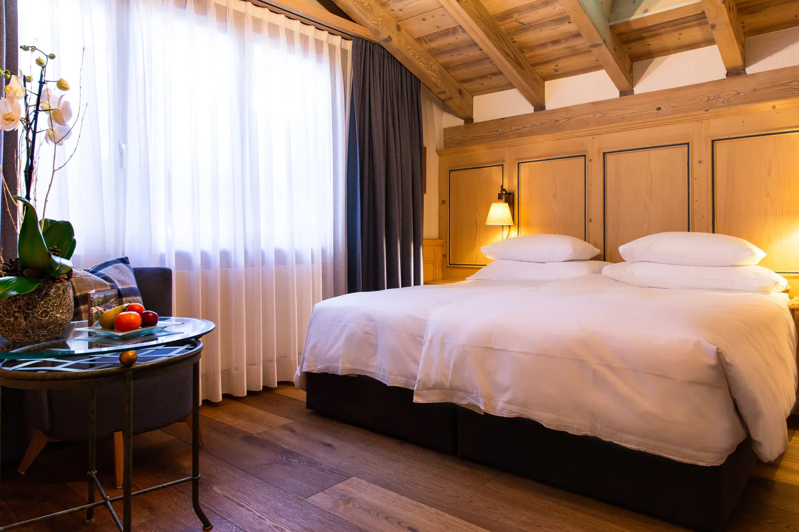 A Plani Compact room: a double bed with white linen under an exposed timber ceiling, a round glass table with fruit, an orchid and a window with long curtains