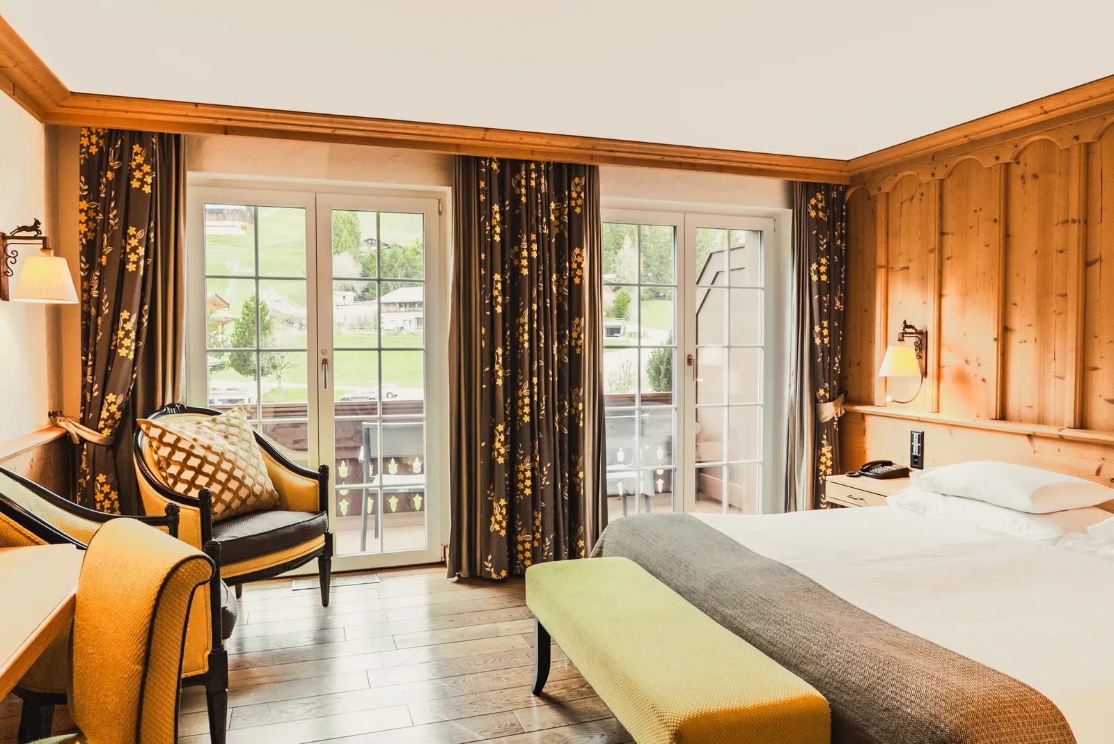 A Rellerli Classic room in the Chalet Saanen: a double bed with a yellow bench at its foot, a mustard armchair, pine panelling, wall lamps and French windows onto a balcony