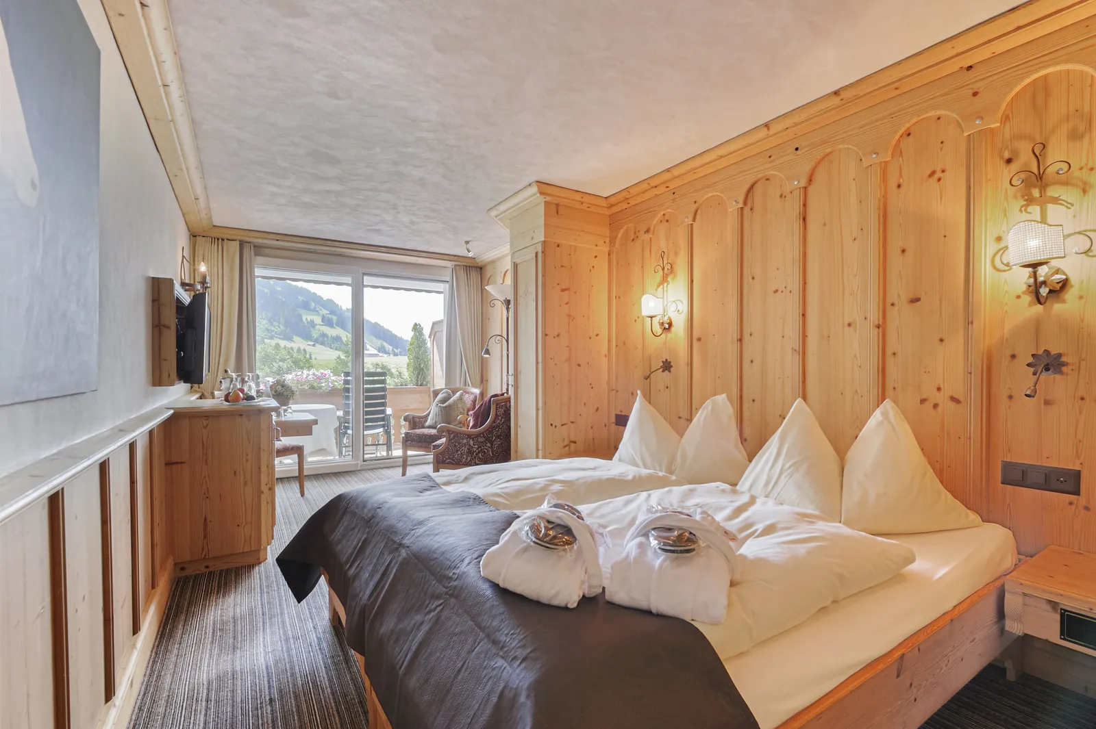 A Ried Superior room: a double bed with white bathrobes, pine panelling with arched motifs, wall lamps, a seating area and a balcony door at the far end