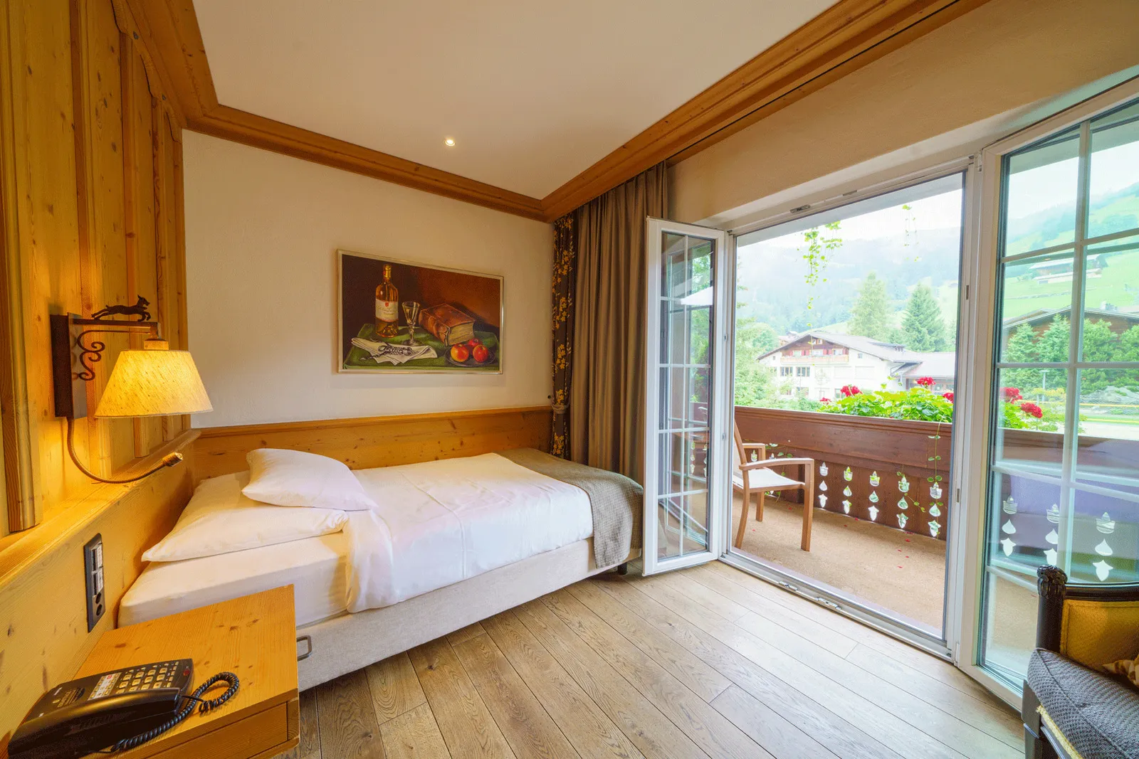 A Hugeli single room: the bed against a wood-panelled wall, a wall lamp, a painting and an open balcony door with red geraniums, meadows and chalets outside