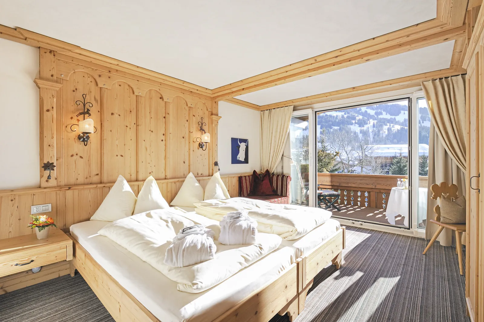 The Eggli Suite bedroom: a double bed with bathrobes, pine panelling with carved details, a carpeted floor and a balcony door onto snow-covered hills