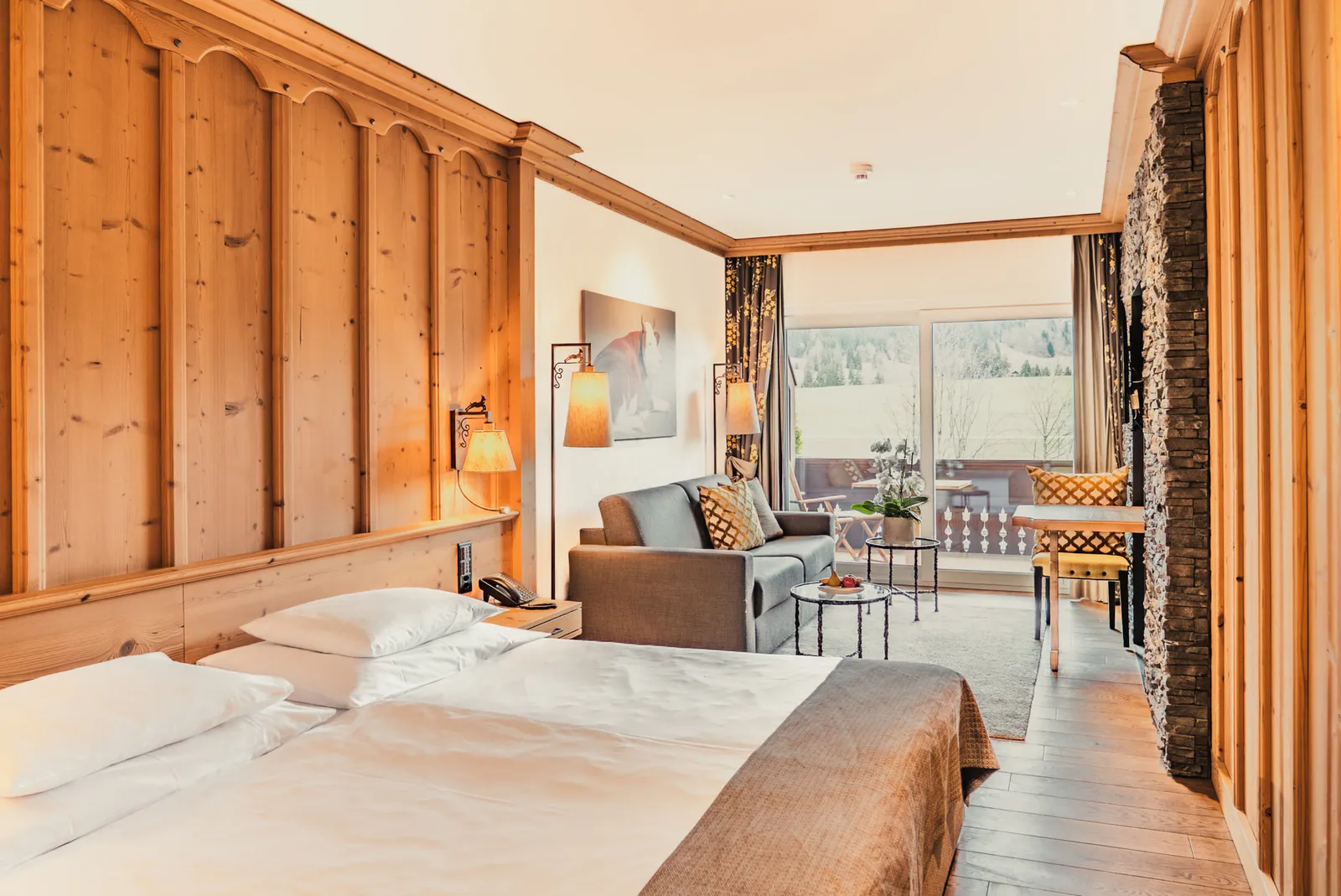 A Gstaad Junior Suite in the Chalet Saane: a double bed, tall pine panelling, a stone wall and, at the far end, a sofa and a table in front of the window onto the garden