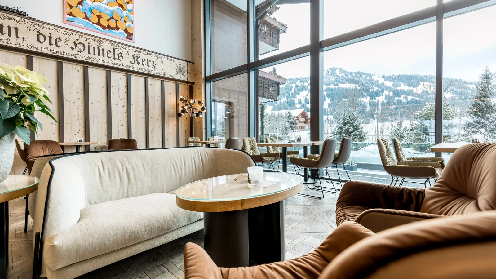The Bärgblick lounge in winter: a cream sofa and brown leather armchairs around a round table, tall windows onto snowy slopes and chalets, a painting of stacked logs in the snow and a painted inscription along the wall