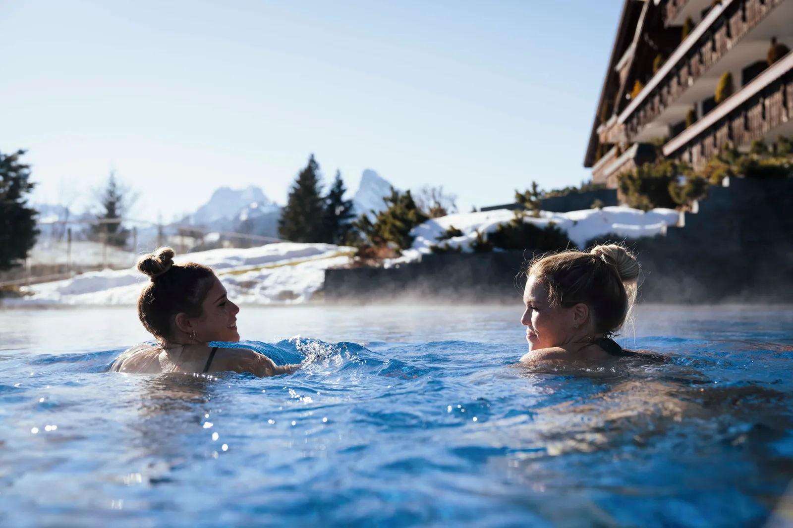 2 women swimming in the steaming outdoor pool, with the chalet balconies on the right and a snowy rocky peak and fir trees behind
