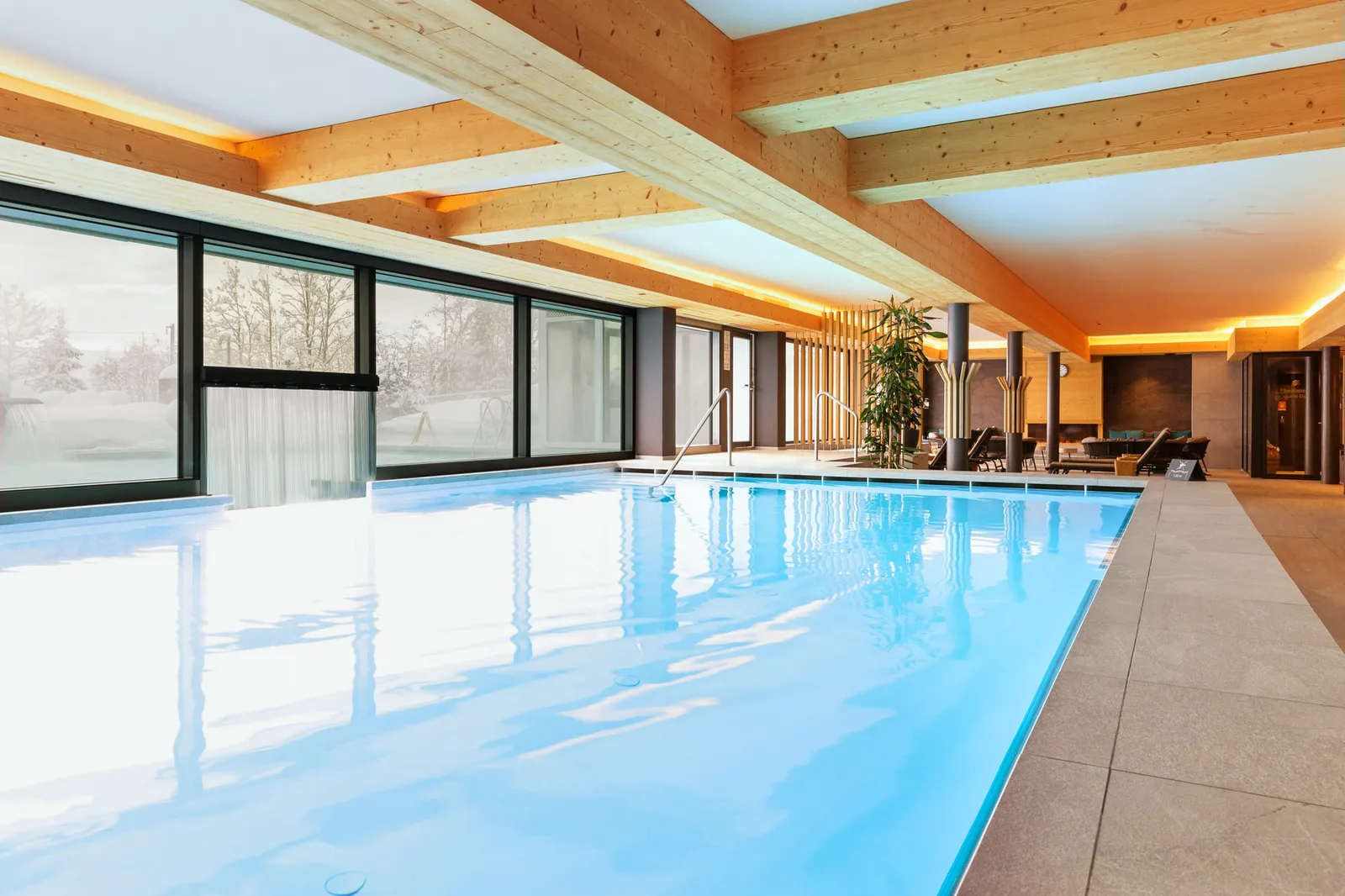 The long indoor brine pool under timber beams and warm strips of light, with windows onto the snow and loungers along the far side