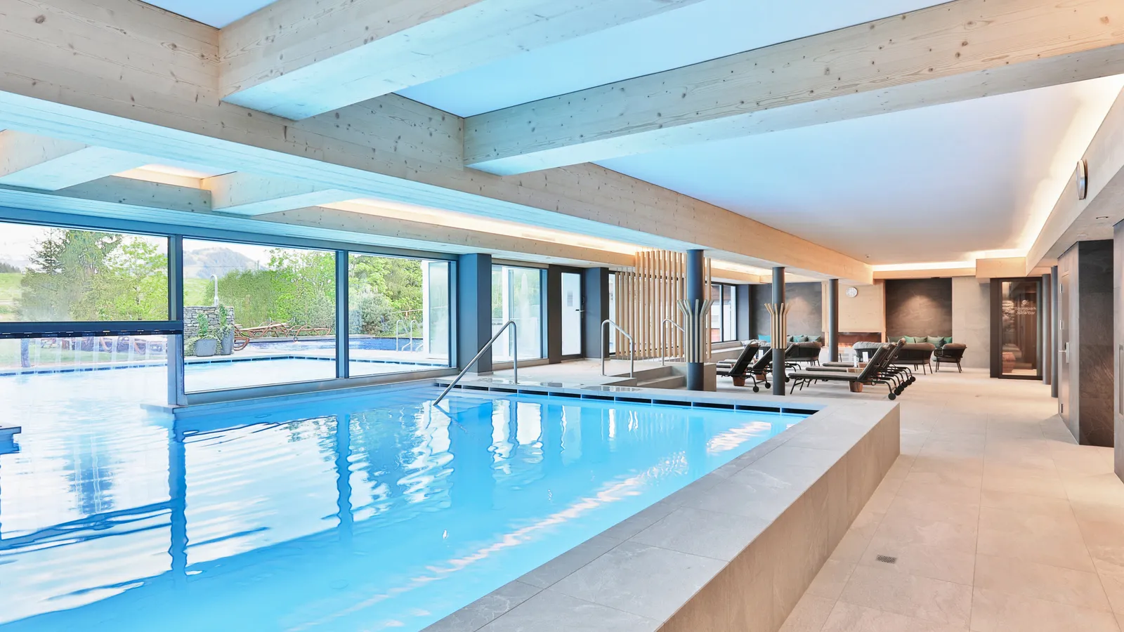 The indoor brine pool under pale timber beams, with the outdoor pool beyond the glass wall, loungers along the far side and spring green outside