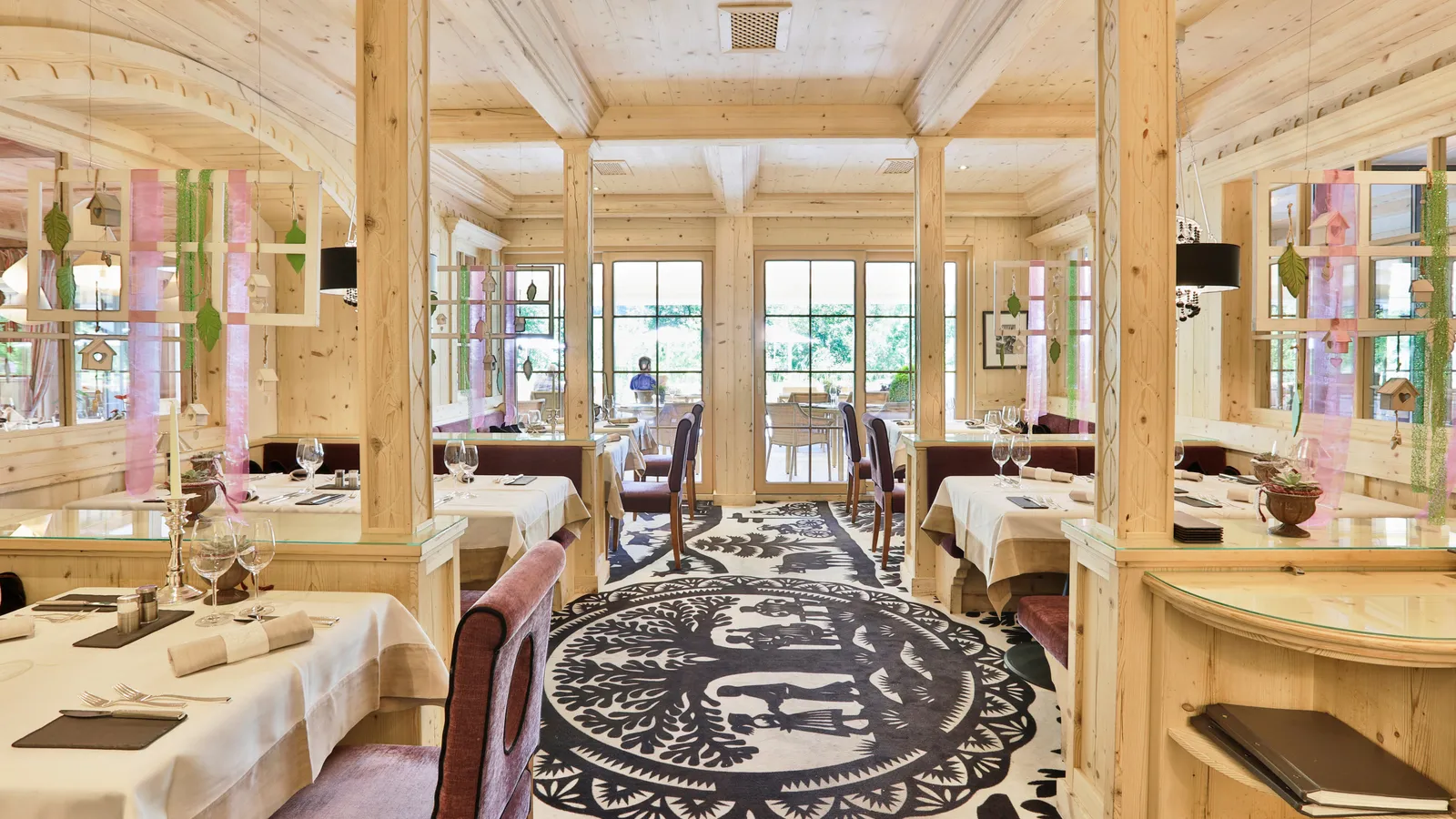 The Ermitage Stube: a dining room in pale pine with carved pillars, tables laid in white and cream, mauve chairs, a white carpet with black papercut figures and French windows onto the terrace