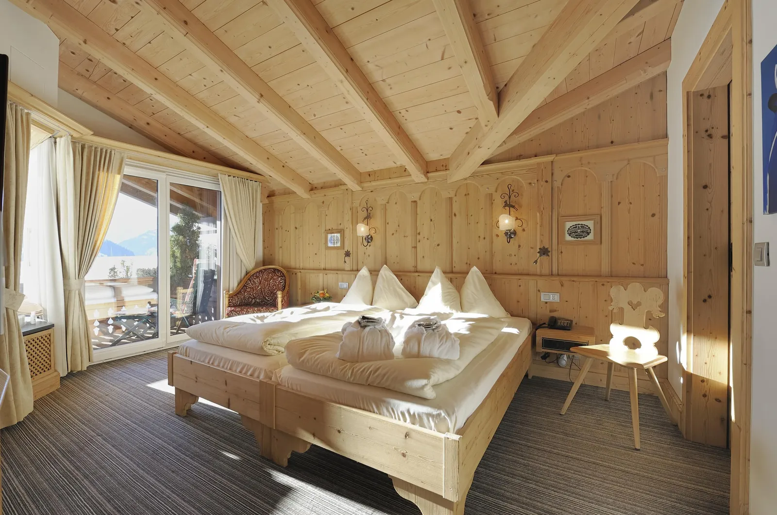 The Wellness Suite bedroom: 2 beds under exposed roof beams, pine panelling, a carved chair, a striped carpet and a balcony door onto the snow