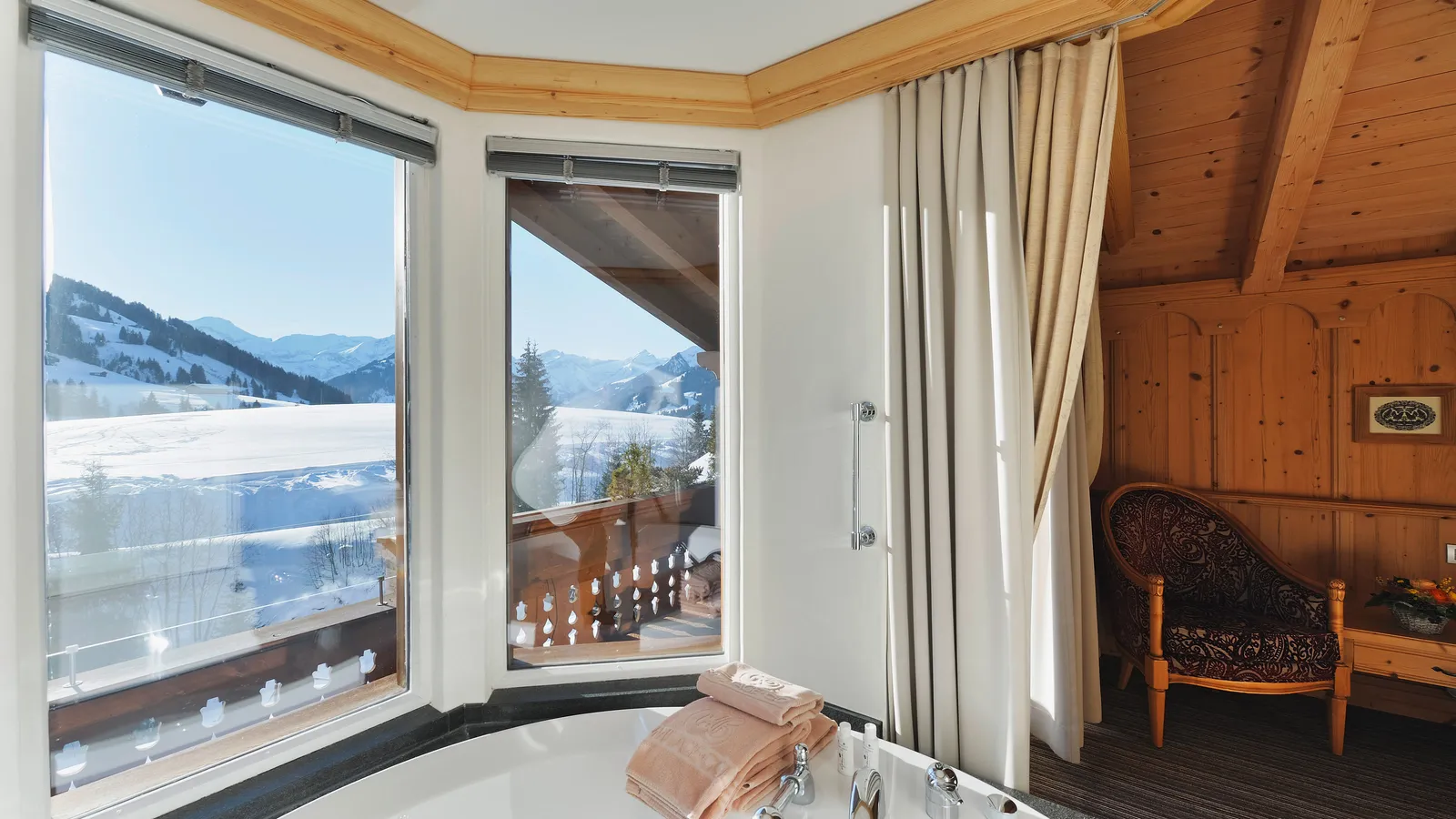 The round whirlpool bath in the Wellness Suite’s glazed bay, with 2 windows onto a snow-covered valley and mountains, a carved balcony railing and a wood-panelled corner with an armchair