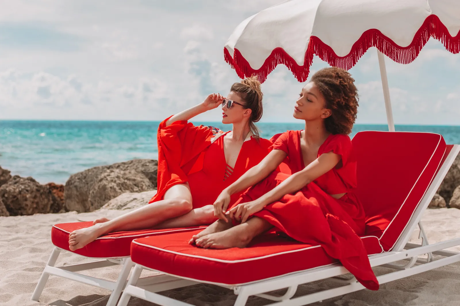 2 women dressed in red on red loungers under a striped umbrella on the sand, the ocean behind them.