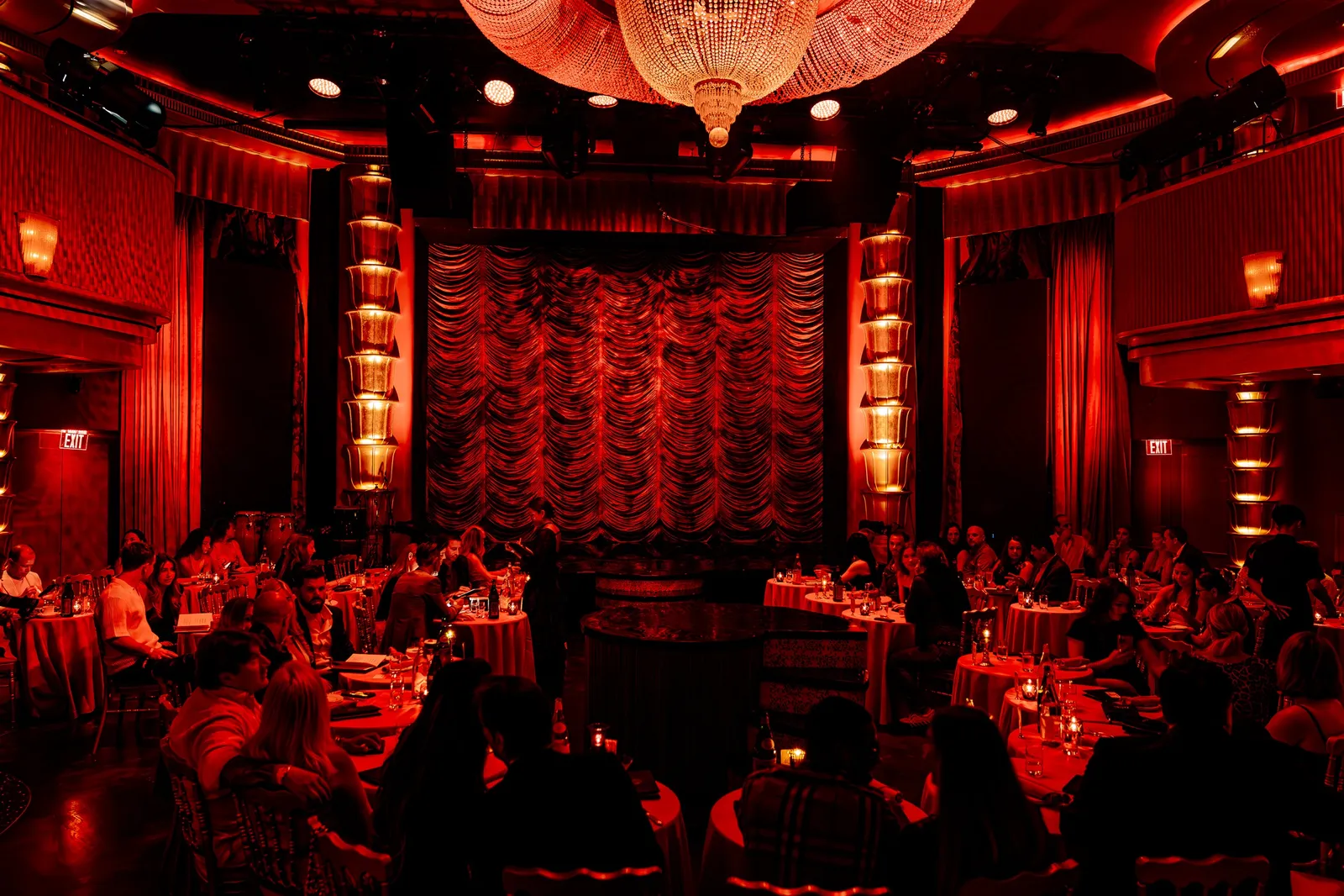 The Faena Theater in red light, the audience at tables before the curtain and a chandelier overhead.