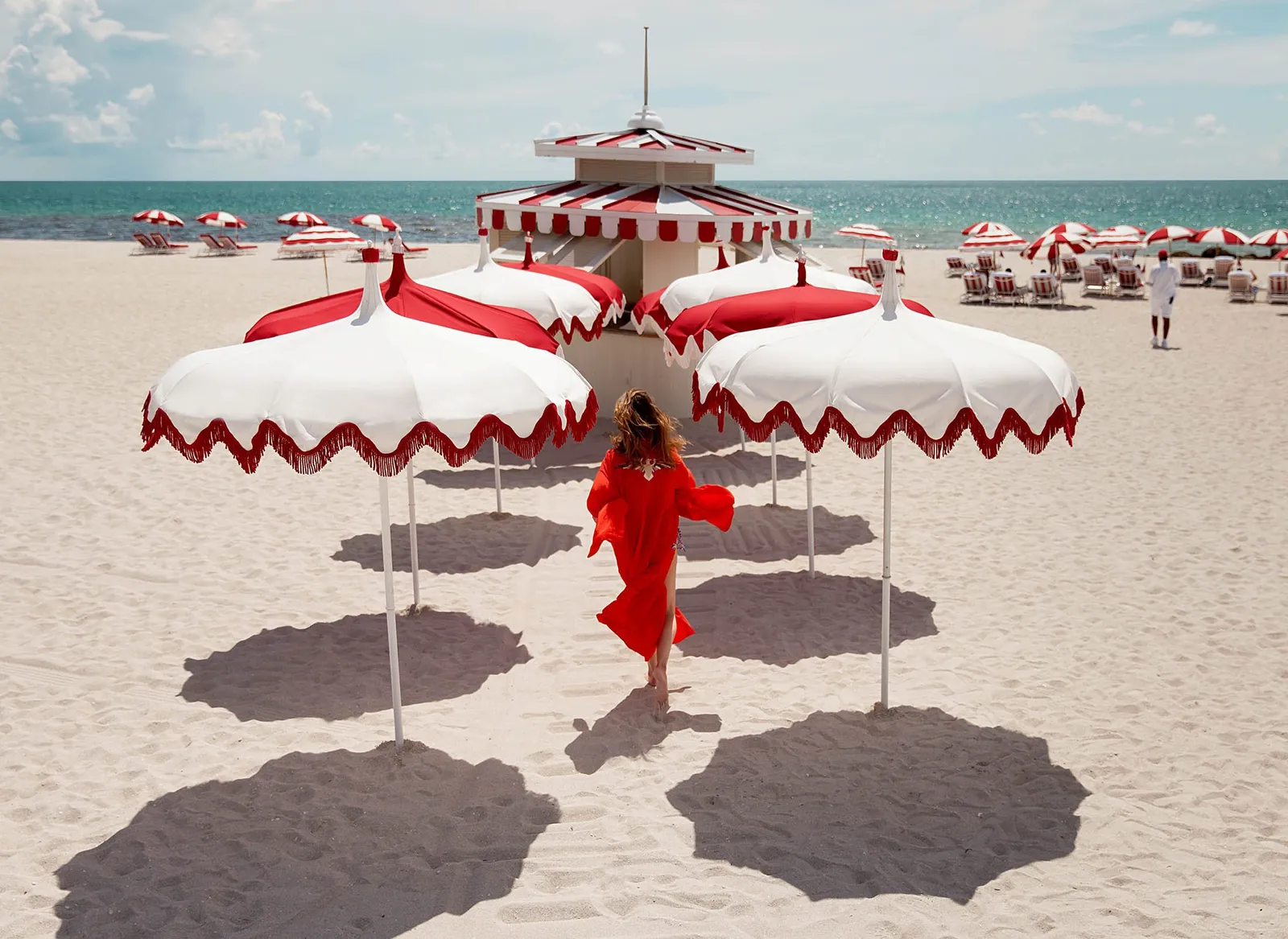 A woman in a red dress walks across the sand among red-and-white umbrellas, the ocean behind her.