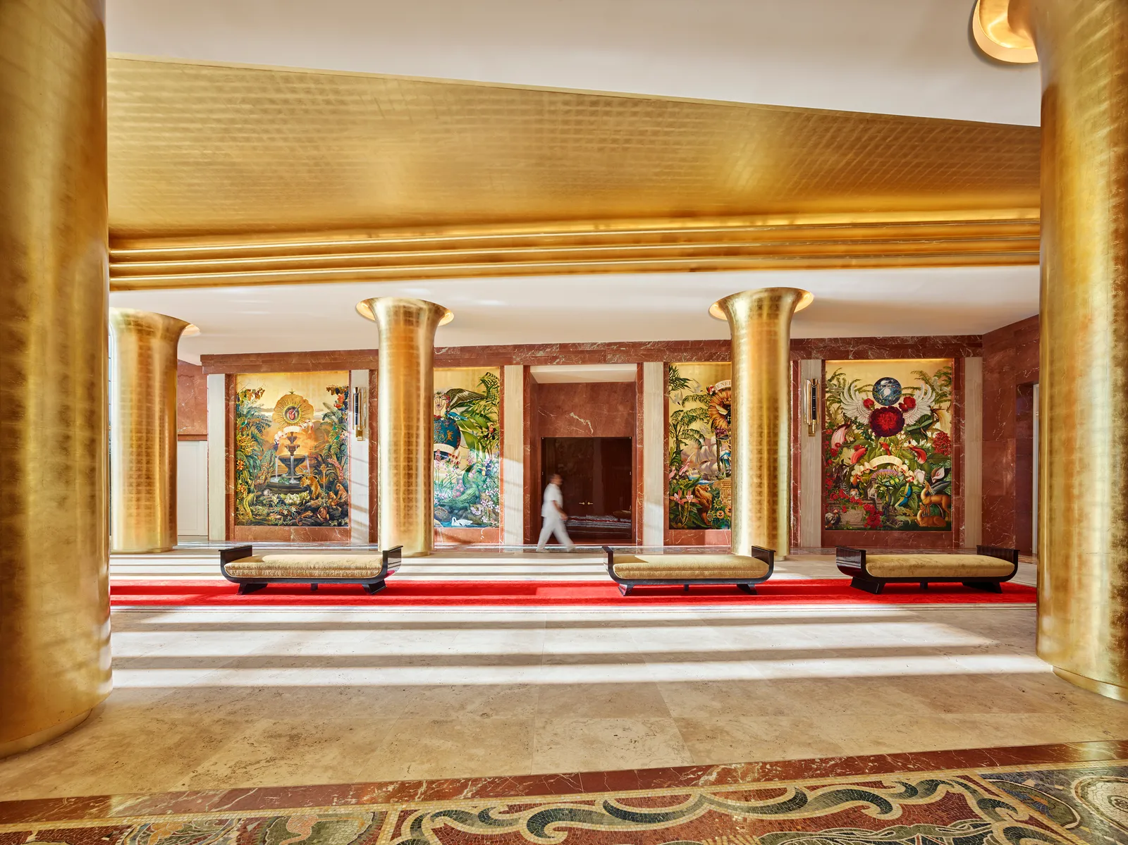 The entrance hall with its gold-leaf columns, murals at the far end, a red carpet and a man walking through.