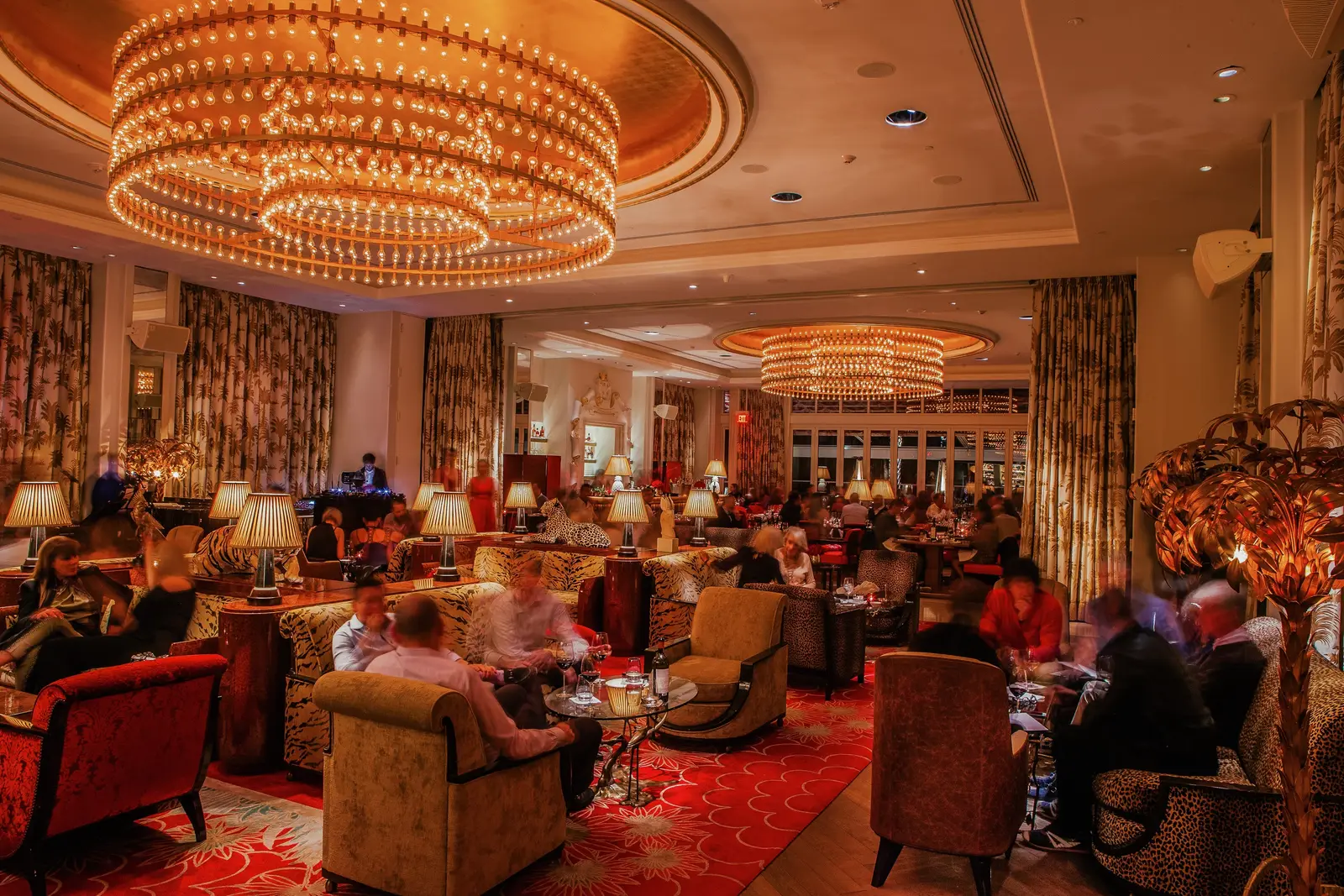 The Living Room in the evening: a great round chandelier, armchairs and sofas with guests seated, and red carpets.