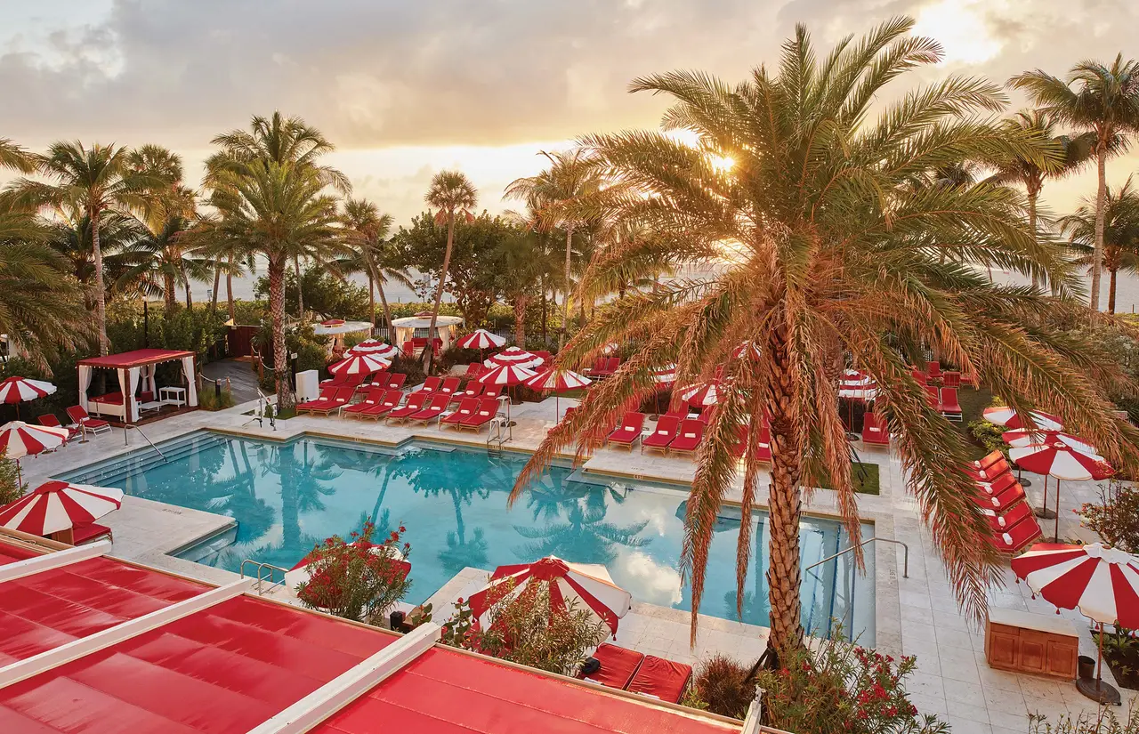 The pool at sunset, ringed by palms, red loungers and striped umbrellas.