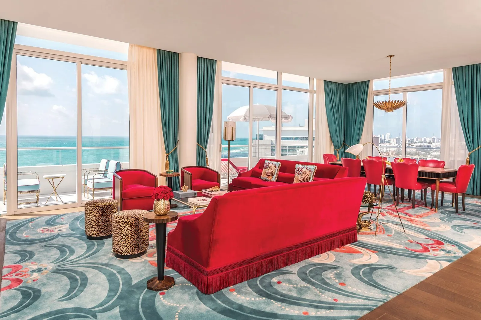 The Oceanfront Three Bedroom Suite’s living room: red sofas on a turquoise carpet, the dining area and windows onto the ocean.