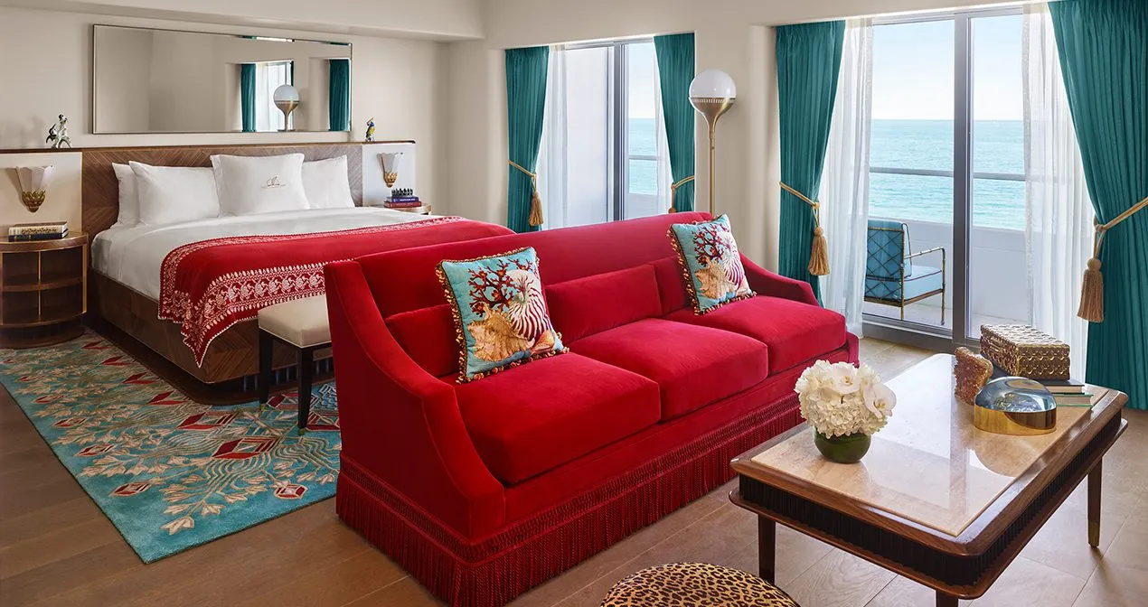 A Premier Oceanfront Room: the bed, the red sofa with flowered cushions, turquoise curtains and the ocean beyond the balcony.