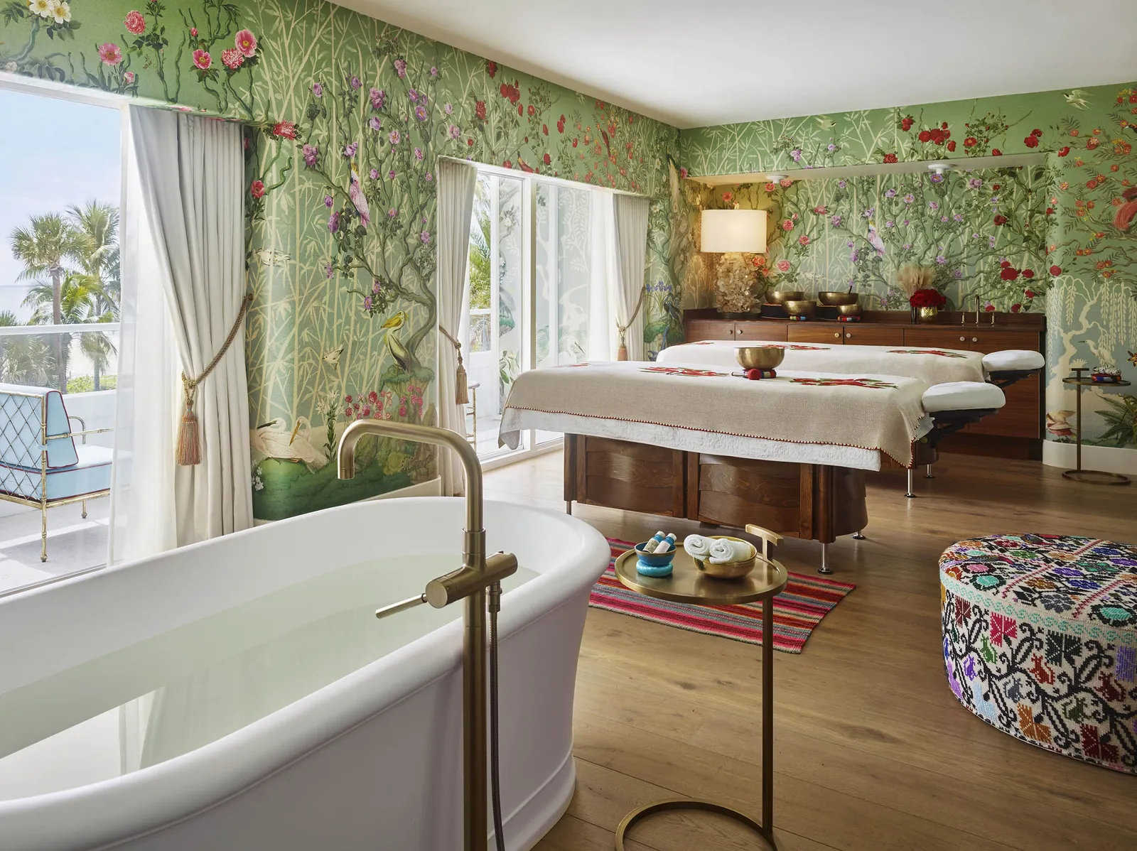 A spa suite: the bathtub in the foreground, 2 treatment beds, flowered wallpaper and a window onto the palms.