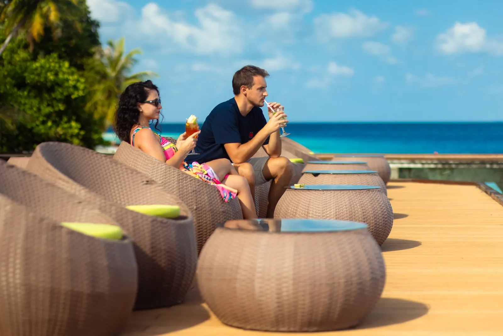A couple drink cocktails in wicker chairs on a timber deck facing the sea.