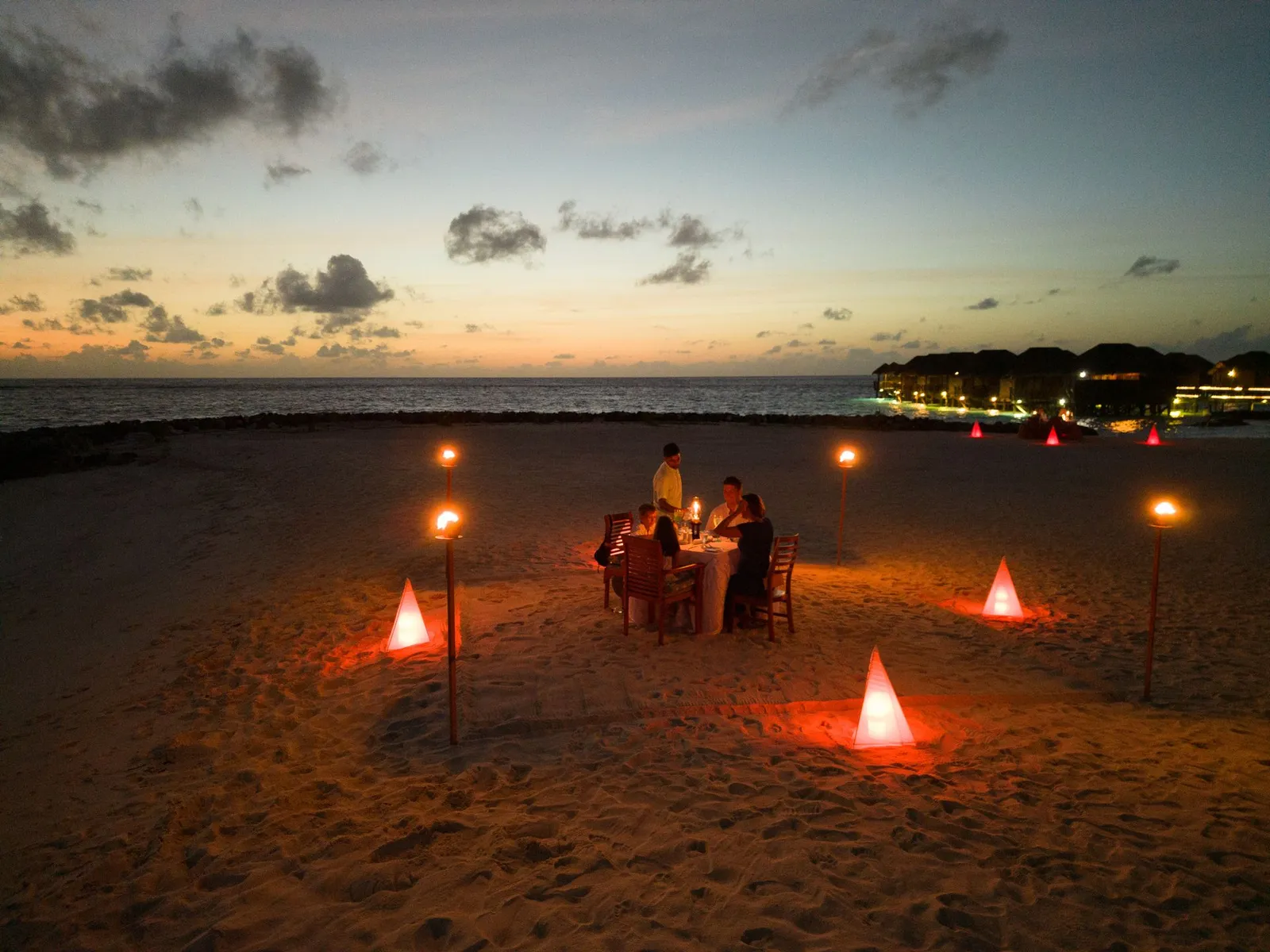 A private dinner on the beach after sunset, among lit torches and lanterns, with the water bungalows’ lights beyond.